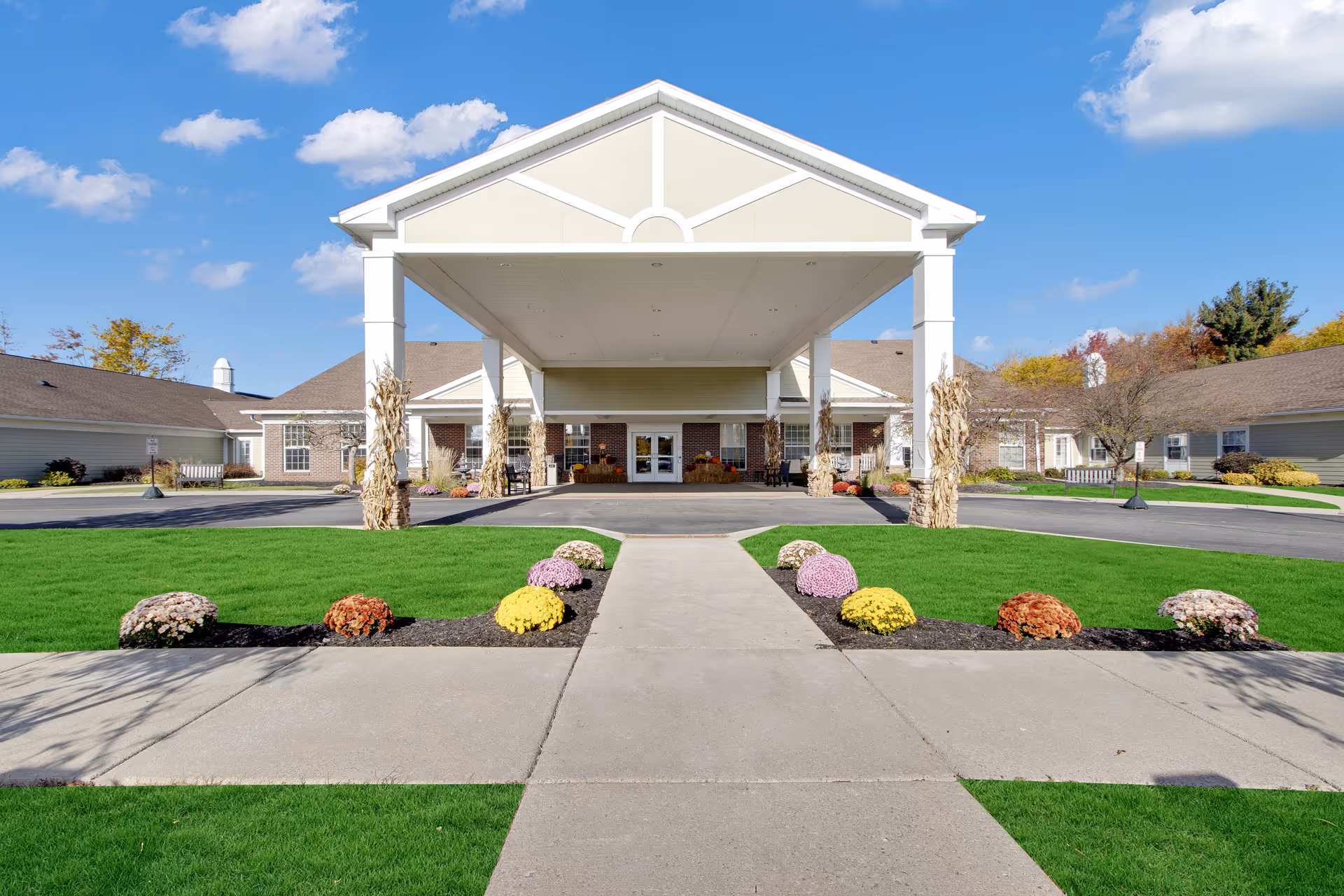 Front exterior view of Orchard Heights senior living facility in Orchard Park, showing a covered entrance with white columns, a paved driveway, green lawns, and flower beds with colorful flowers under a blue sky with some clouds.