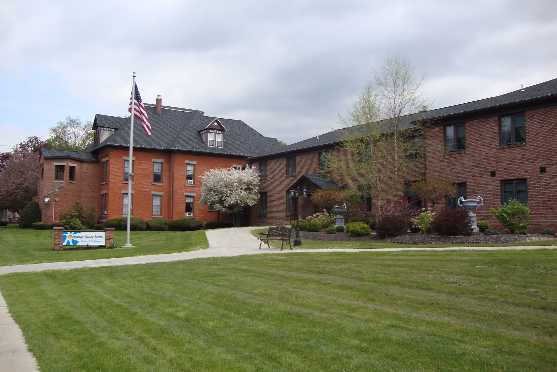 Exterior view of Chenango Valley Home, a brick building with two connected sections, surrounded by a well-maintained lawn, trees, shrubs, and a flagpole with an American flag. A bench and a sign with the facility name are visible near the entrance.