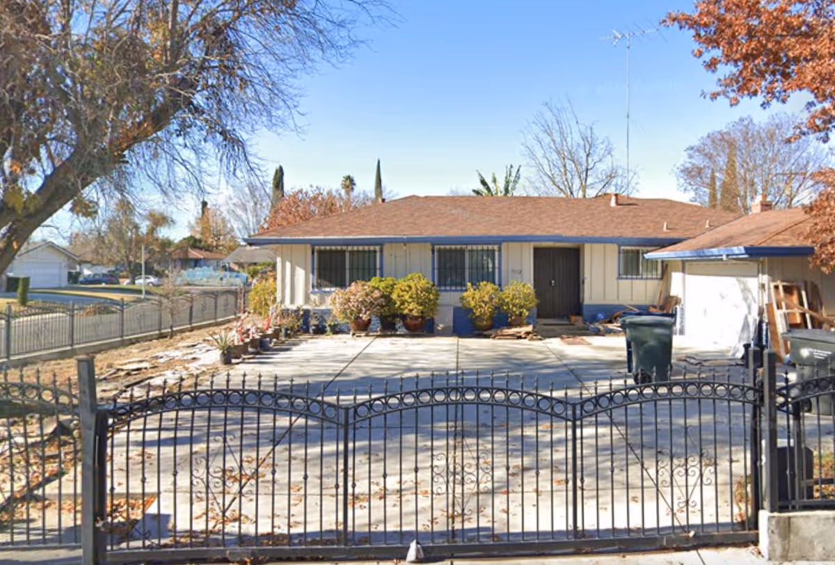Front exterior view of a single-story house with a brown roof, white walls, and blue trim. There is a black wrought iron gate and fence in the foreground, a concrete driveway, several potted plants near the house, and two green trash bins on the right side. Trees with autumn leaves surround the property.