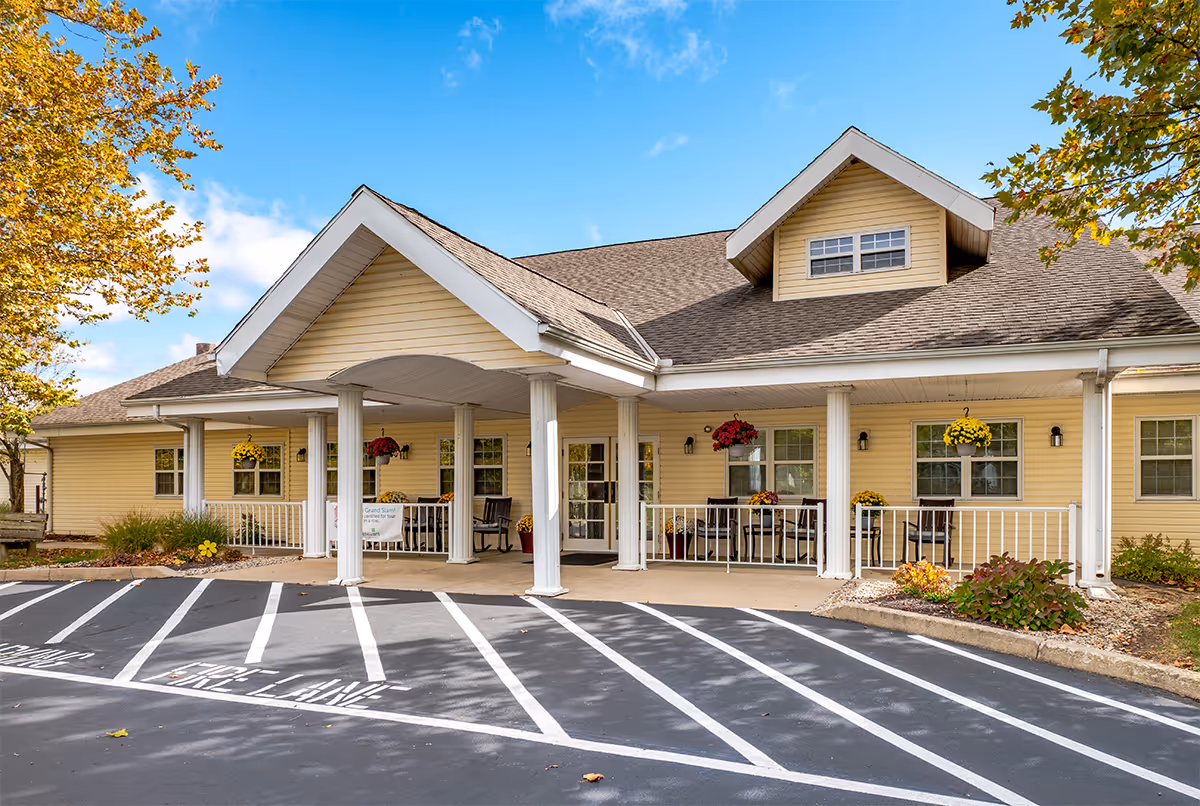 Exterior front view of a single-story yellow building with a covered entrance supported by white columns. There are hanging flower baskets and chairs on the porch. The parking area in front has marked fire lane lines. Trees with autumn leaves frame the scene under a blue sky with some clouds.