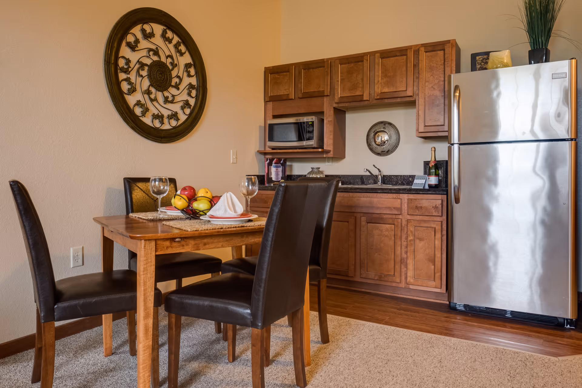 A small kitchen and dining area featuring wooden cabinets, a stainless steel refrigerator, a microwave, and a sink. A wooden dining table with four black leather chairs is set with plates, napkins, wine glasses, and a fruit basket. A decorative circular wall art piece hangs on the wall.