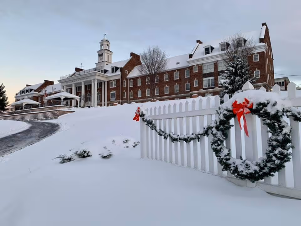 A large brick building with white columns and a cupola, covered in snow. In the foreground, there is a white picket fence decorated with a snow-covered green garland and a wreath with a red bow. The ground and trees are also covered in snow, indicating a winter scene.