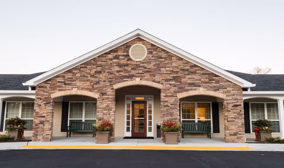 Front exterior view of a senior living facility with a stone facade, three arched openings, two green benches, potted plants with red flowers, and a central glass door entrance.