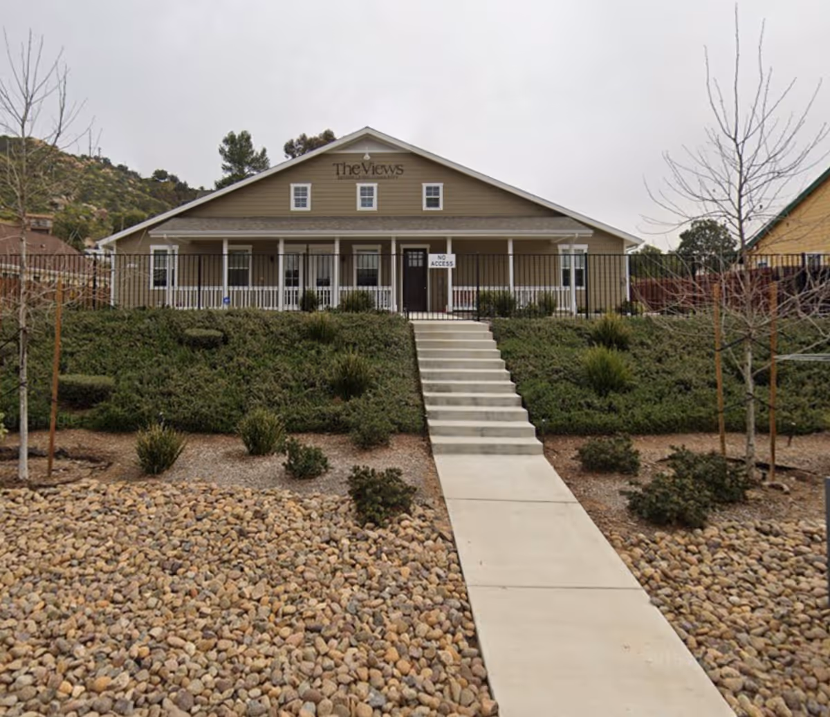 Front exterior view of a single-story building with a gable roof, beige siding, and a porch with white railings. A concrete walkway and stairs lead up to the entrance, which has a 'No Access' sign. The surrounding landscape includes rocks, shrubs, and leafless trees under an overcast sky.