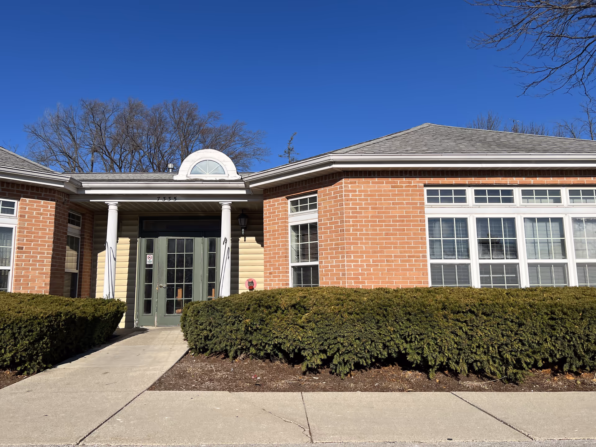 Exterior front view of a single-story brick building with large windows and a central entrance featuring double glass doors and white columns. There are trimmed bushes along the front and a clear blue sky above.
