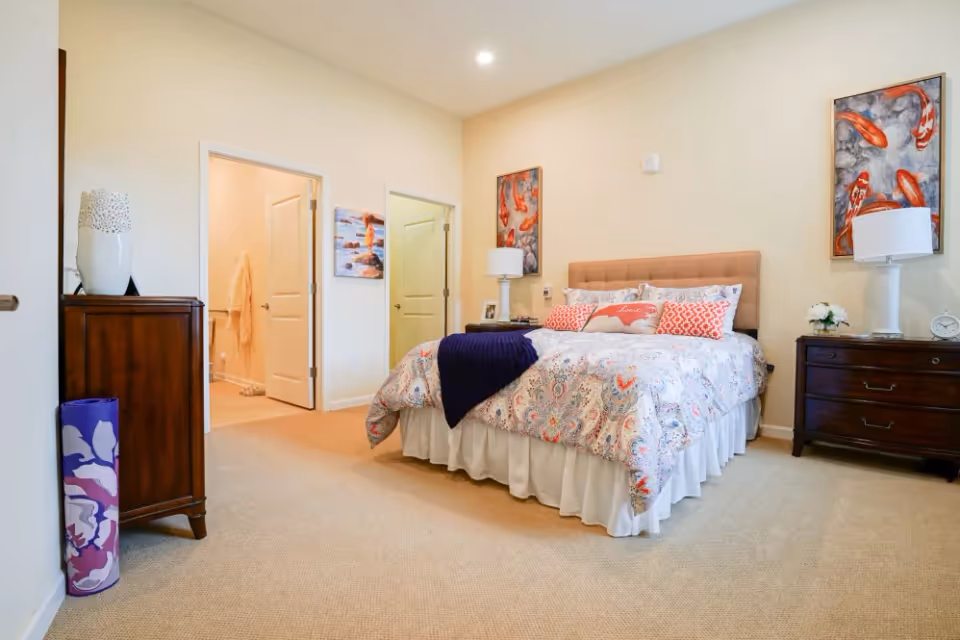 A neatly arranged bedroom with a large bed covered in a patterned bedspread and several pillows, including one with the word 'home'. The room has beige walls and carpet, two dark wooden nightstands with white lamps, and colorful artwork featuring koi fish on the walls. There are two open doors leading to other rooms, one of which appears to be a bathroom with a robe hanging inside. A rolled-up yoga mat is placed near a wooden dresser on the left side.