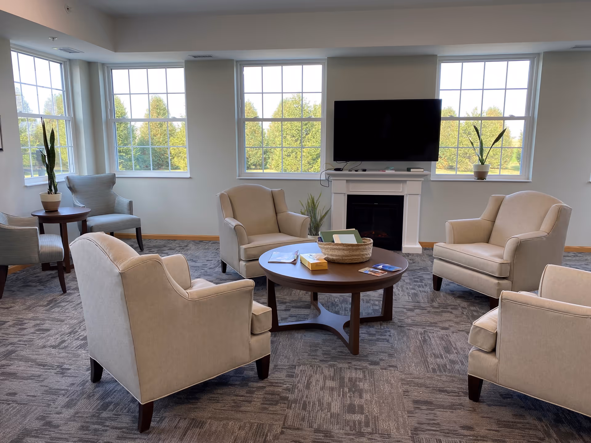 A bright and spacious living room area with four beige armchairs arranged around a round wooden coffee table. The table has a basket, some books, and pamphlets on it. Behind the seating area is a white fireplace with a flat-screen TV mounted above it. Large windows let in natural light and offer a view of green trees outside. Two potted plants are placed on side tables near the windows.