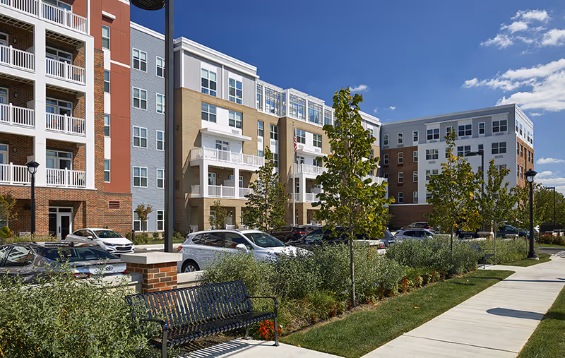 Exterior view of a senior living facility with multi-story buildings featuring balconies, a parking lot with cars, a sidewalk, benches, and landscaped greenery under a blue sky with some clouds.