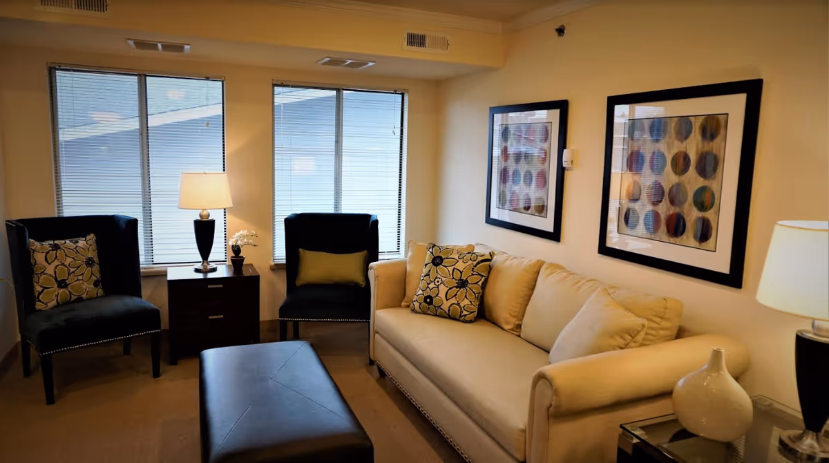 A cozy living room with a beige sofa adorned with floral cushions, two dark armchairs with patterned pillows, a dark ottoman in the center, two framed abstract artworks on the wall, and two table lamps on side tables. Large windows with blinds allow natural light into the room.