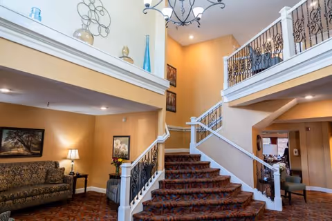 Interior view of a senior living facility lobby with a carpeted staircase featuring ornate black railings. To the left, there is a sitting area with a patterned sofa, a side table with a lamp, and framed artwork on the walls. The walls are painted a warm beige color, and decorative vases and wall art are displayed on a ledge above the seating area. A chandelier hangs from the ceiling, illuminating the space.