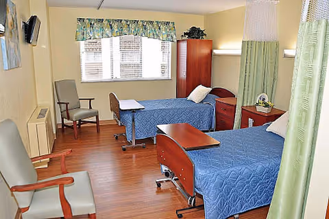 A well-lit room with two hospital-style beds covered with blue quilts, each with a pillow and a wooden overbed table. There are two chairs with wooden armrests, a wooden cabinet, a window with blinds and a floral valance, and green privacy curtains partially drawn around the beds. The floor is wooden, and the walls are painted light yellow.
