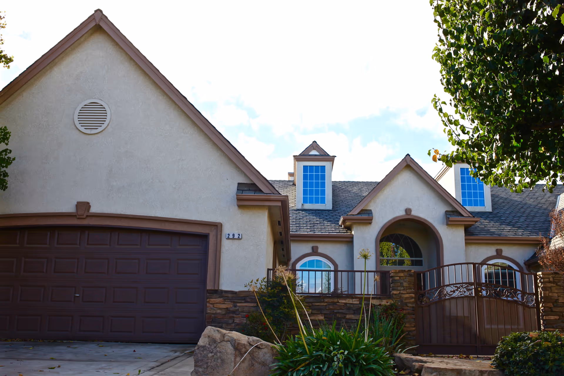 Exterior view of a residential-style building with beige stucco walls, brown trim, and a dark brown garage door. The building has multiple peaked roofs with small dormer windows. There is a gated entrance with stone pillars and greenery including bushes and a tree in the foreground.