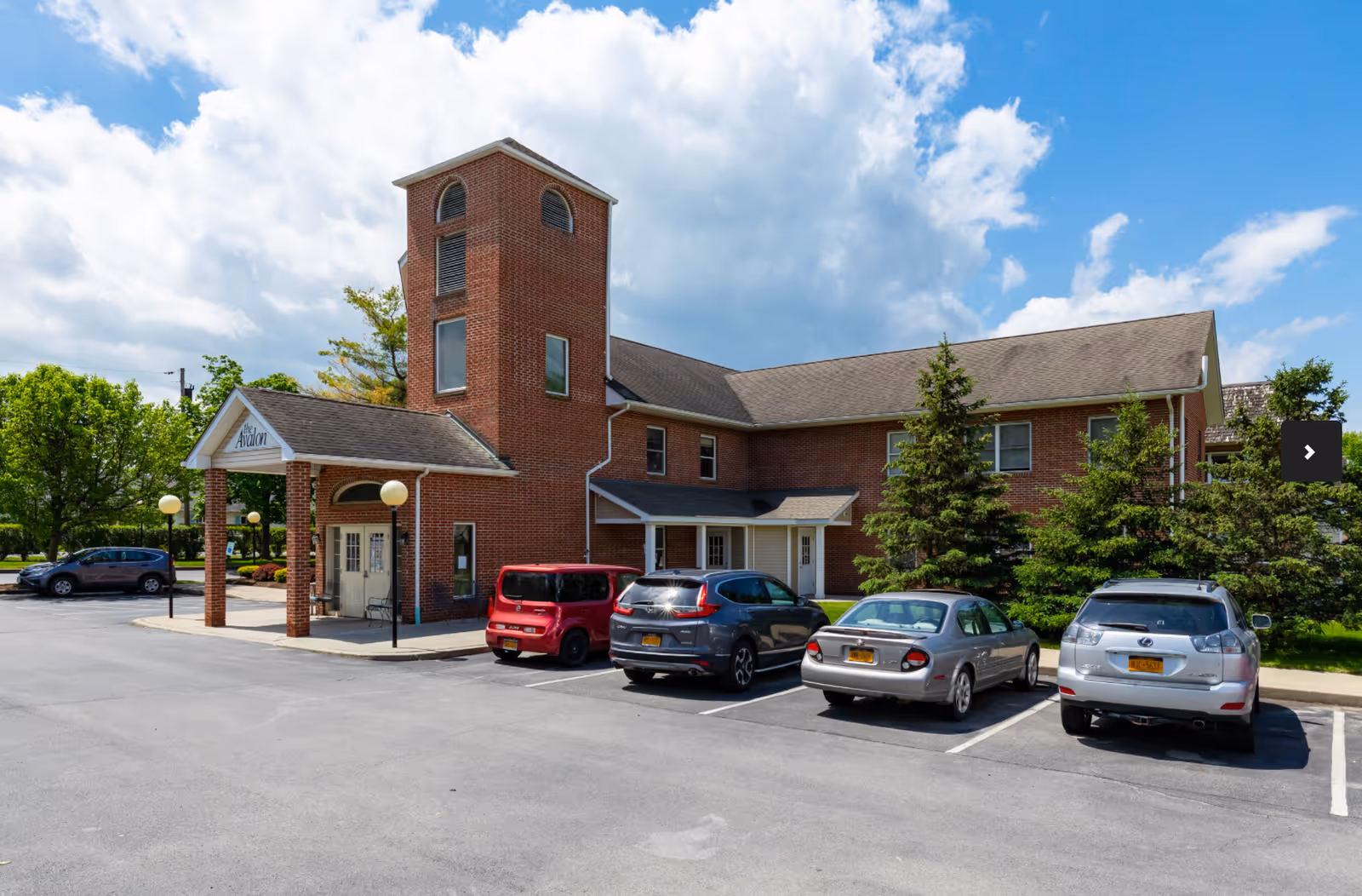 Exterior view of Avalon Assisted Living facility showing a two-story brick building with a tower-like structure, a covered entrance, several parked cars, and trees under a partly cloudy blue sky.