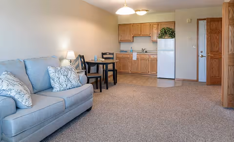A cozy living area with a light blue sofa adorned with two patterned pillows, a small round dining table with two chairs, and a kitchenette featuring wooden cabinets, a white refrigerator, and a sink. The room has beige carpeting and neutral-colored walls with two ceiling lights.