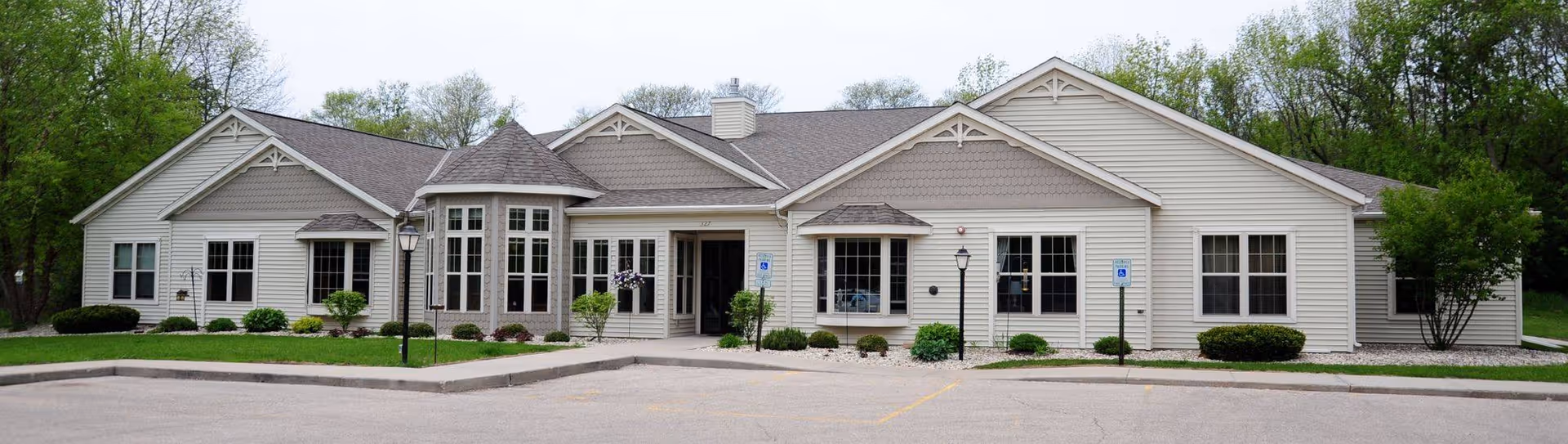 Front exterior view of a single-story senior living facility building with beige siding, multiple windows, and a central entrance. The building is surrounded by small shrubs and trees, with a paved parking area in front including handicap parking spaces.
