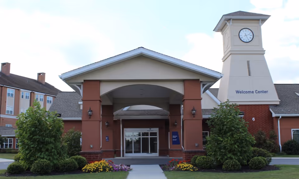 Front exterior view of Brethren Village Village Manor showing the main entrance with a covered driveway and a clock tower labeled 'Welcome Center'. There are bushes and flower beds along the walkway leading to the entrance.