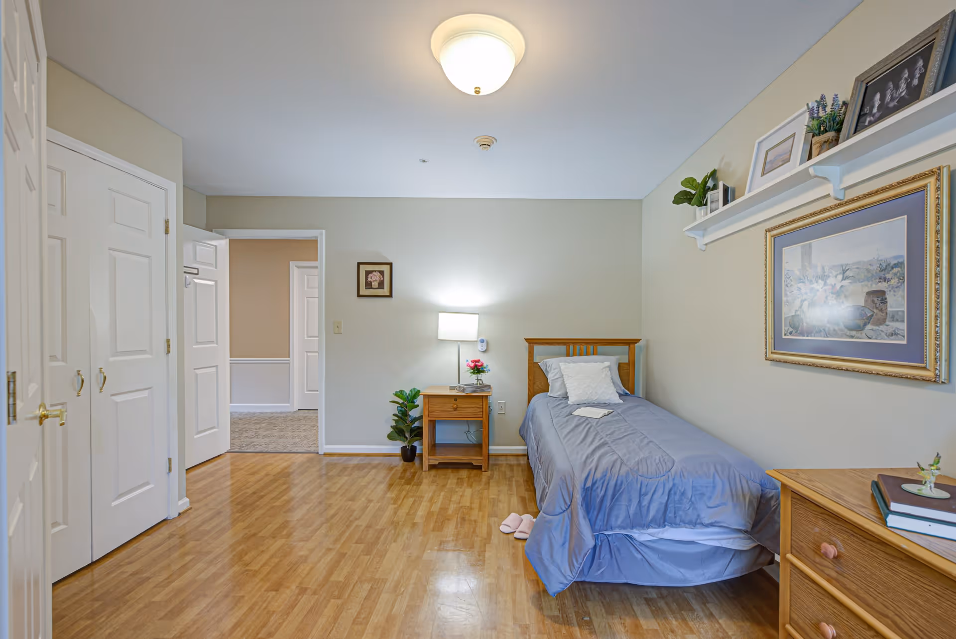 A simple, clean bedroom with a single bed covered in a blue comforter and a white pillow. Next to the bed is a wooden nightstand with a lamp, a small plant, and a flower vase. The room has light-colored walls with framed artwork and decorative shelves holding plants and picture frames. There is a wooden dresser on the right side and a closet with white doors on the left. The floor is wooden, and the room is well-lit with a ceiling light.