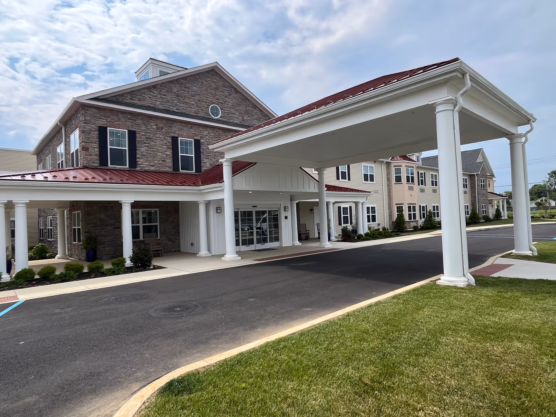 Exterior view of The Birches of Lehigh Valley senior living facility showing a large covered entrance with white columns, a stone and beige siding building with multiple windows, a paved driveway, and a grassy lawn under a partly cloudy sky.