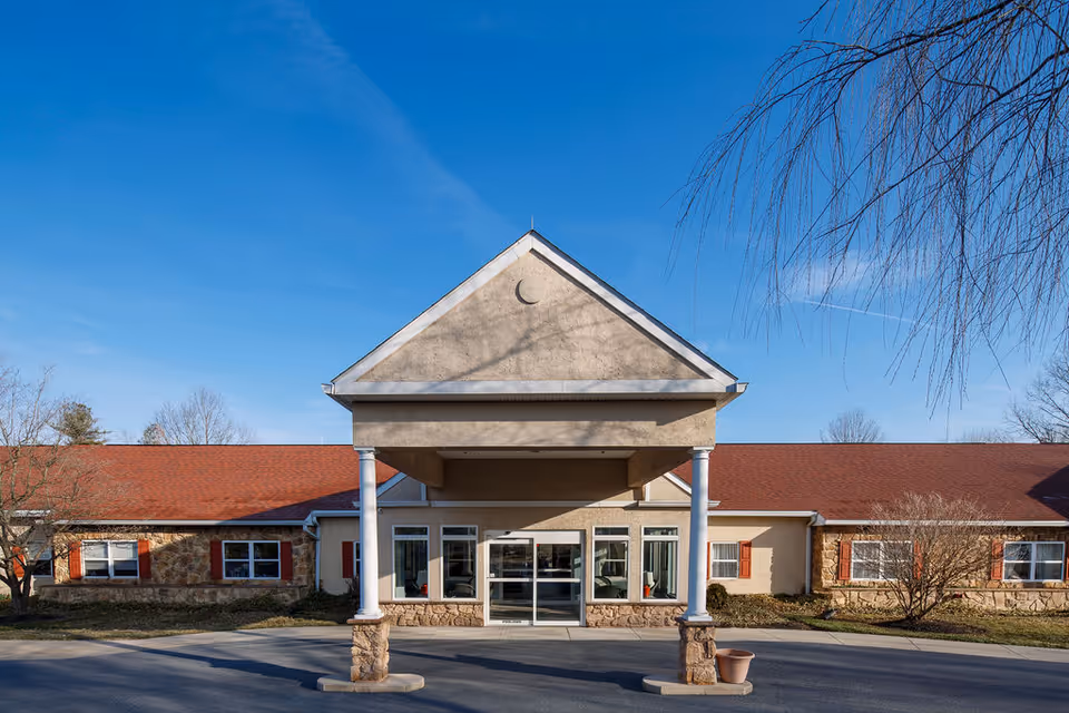 Front exterior view of Oak Leaf Manor South facility with a covered entrance supported by white columns, beige walls, red roof, and windows with red shutters under a clear blue sky.
