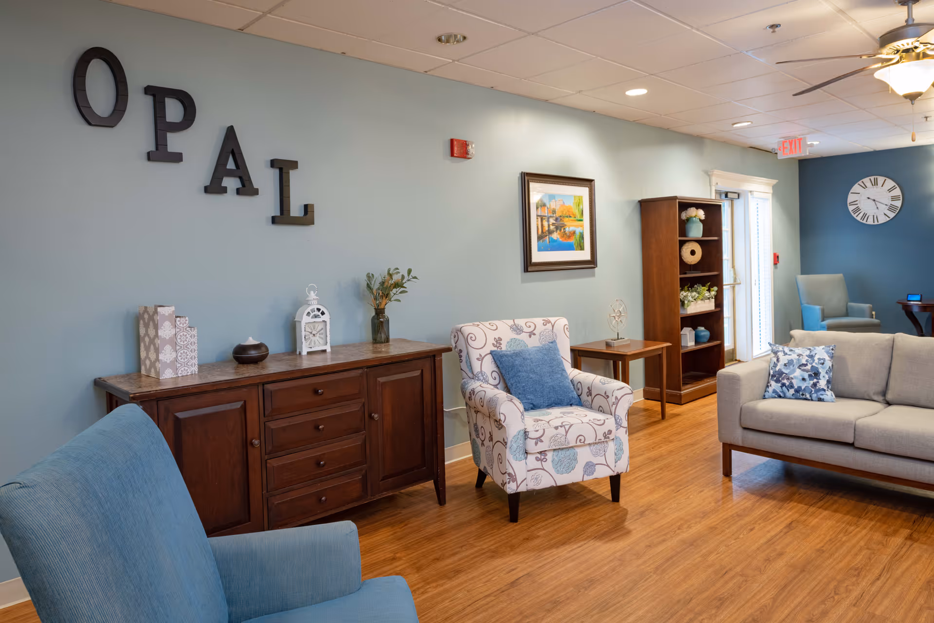 A cozy senior living common area with light blue walls and wooden flooring. The room features a blue armchair, a patterned armchair with a blue cushion, and a light gray sofa with a floral cushion. A wooden sideboard with decorative items, a wooden bookshelf with plants and decor, a framed painting, and a large wall clock are also visible. The word 'OPAL' is displayed on the wall in large black letters.
