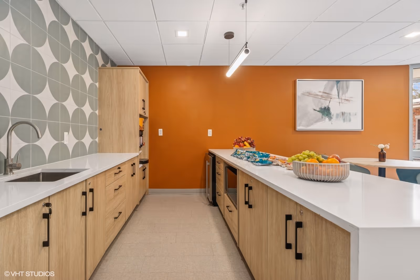 Modern kitchen area with light wood cabinets, white countertops, and a geometric patterned backsplash. A long island counter holds bowls of fruit and snacks. The back wall is painted orange with a framed abstract painting. A pendant light hangs from the ceiling, and there is a window letting in natural light.