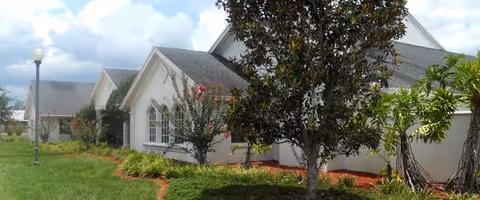 Exterior view of a white building with multiple peaked roofs, surrounded by green grass, trees, and landscaping with red mulch under a partly cloudy sky.