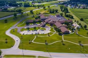 Aerial view of a senior living facility with multiple connected buildings surrounded by green lawns, parking lots with cars, and nearby roads. The facility is situated in a semi-rural area with open fields and scattered trees.