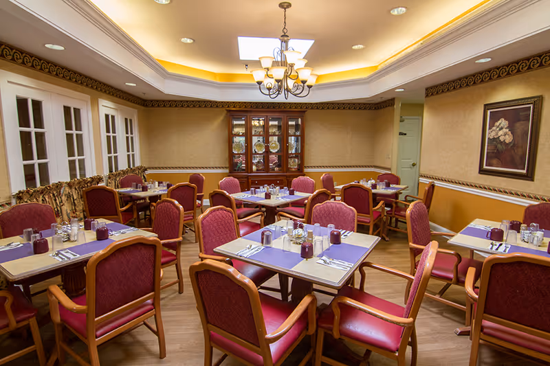 A dining room with multiple tables set with placemats, utensils, glasses, and condiments. The room features red cushioned chairs with wooden frames, a chandelier hanging from the ceiling, a skylight, a china cabinet with decorative plates, and framed artwork on the wall.
