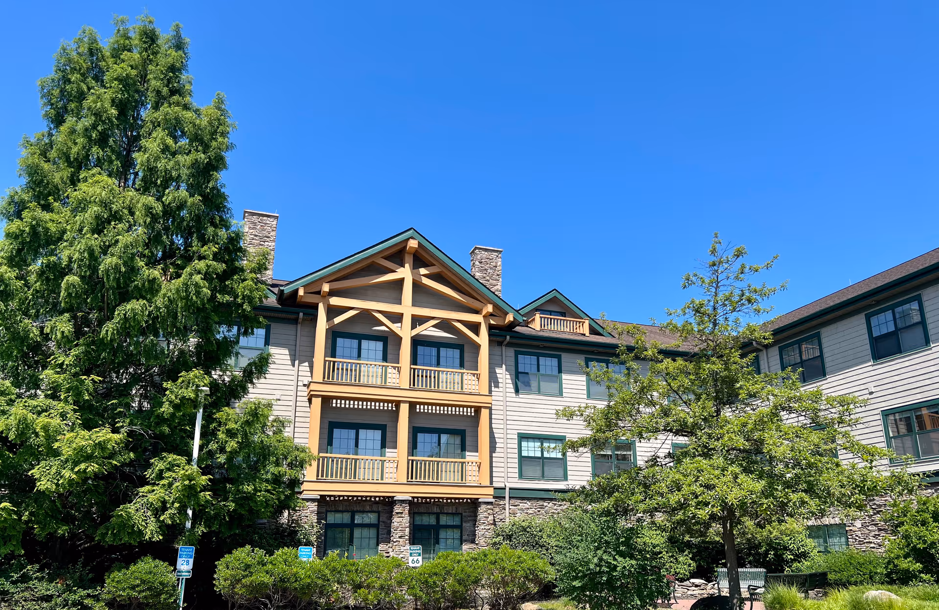 Exterior view of a multi-story senior living facility building with beige siding, green window frames, and wooden balcony railings. The building is surrounded by green trees and shrubs under a clear blue sky.