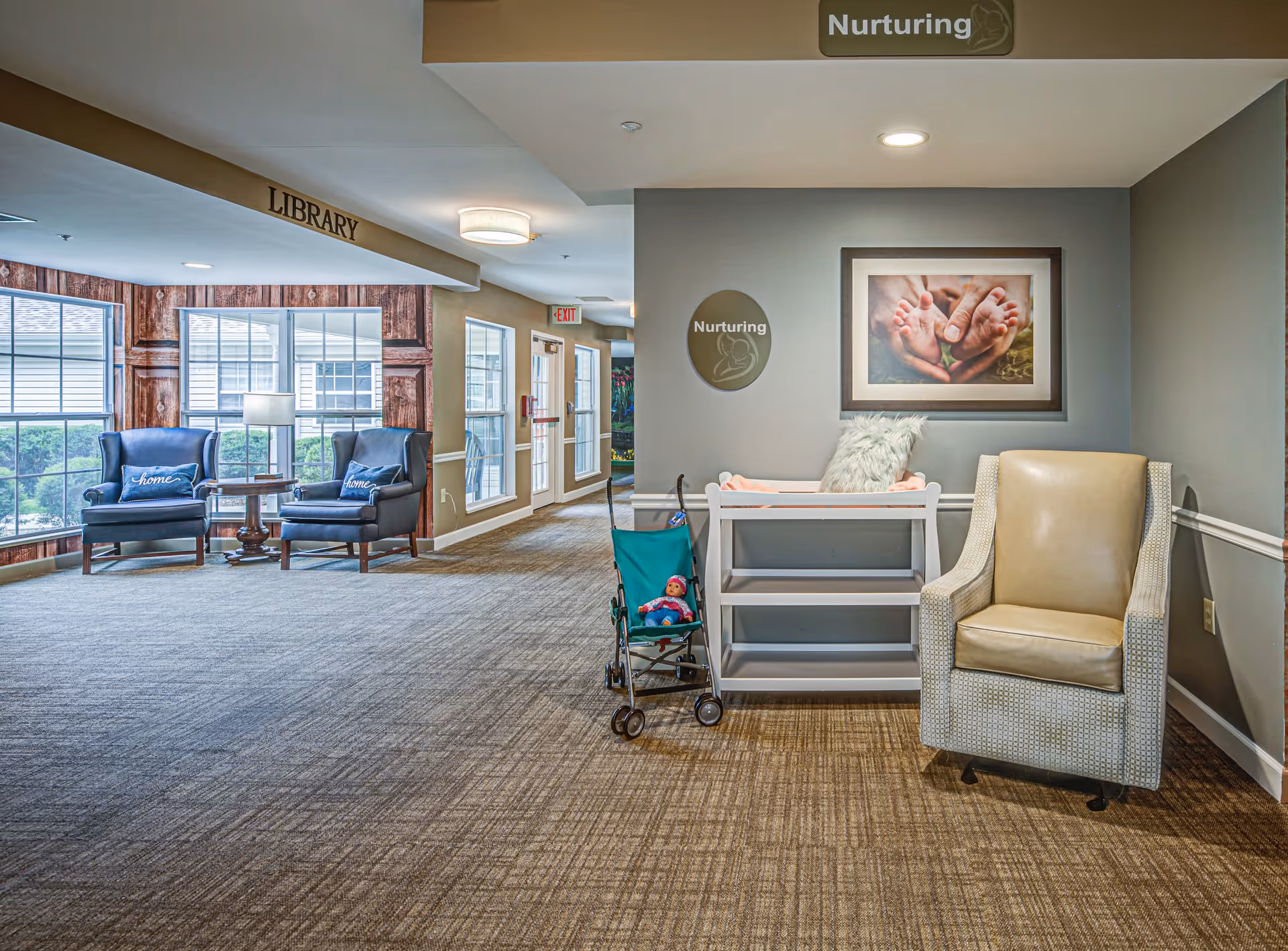 A cozy interior hallway area in a senior living facility with a beige armchair, a small white changing table, and a toy stroller with a doll. On the wall above the changing table is a framed picture of adult hands holding baby feet and two signs that say 'Nurturing.' In the background, there is a seating area labeled 'Library' with two blue armchairs, a small table, and large windows letting in natural light.