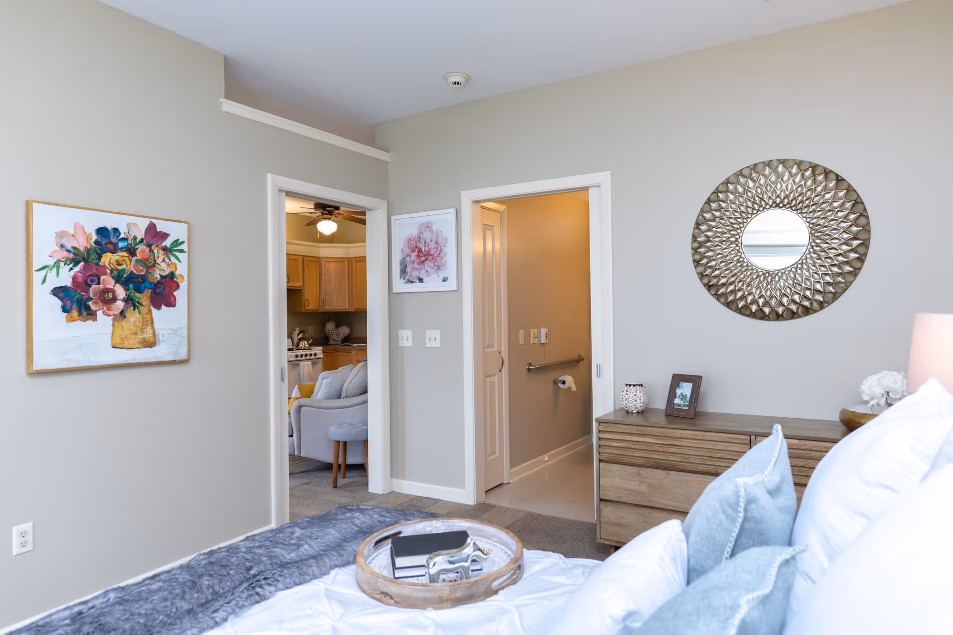 A bedroom with a bed in the foreground featuring white and blue pillows and a gray throw blanket. On the bed is a round wooden tray with books and a decorative item. The room has light gray walls with two framed floral paintings and a decorative round mirror above a wooden dresser. Two doorways are visible, one leading to a kitchen area with wooden cabinets and a ceiling fan, and the other leading to a bathroom with a grab bar and toilet paper holder.