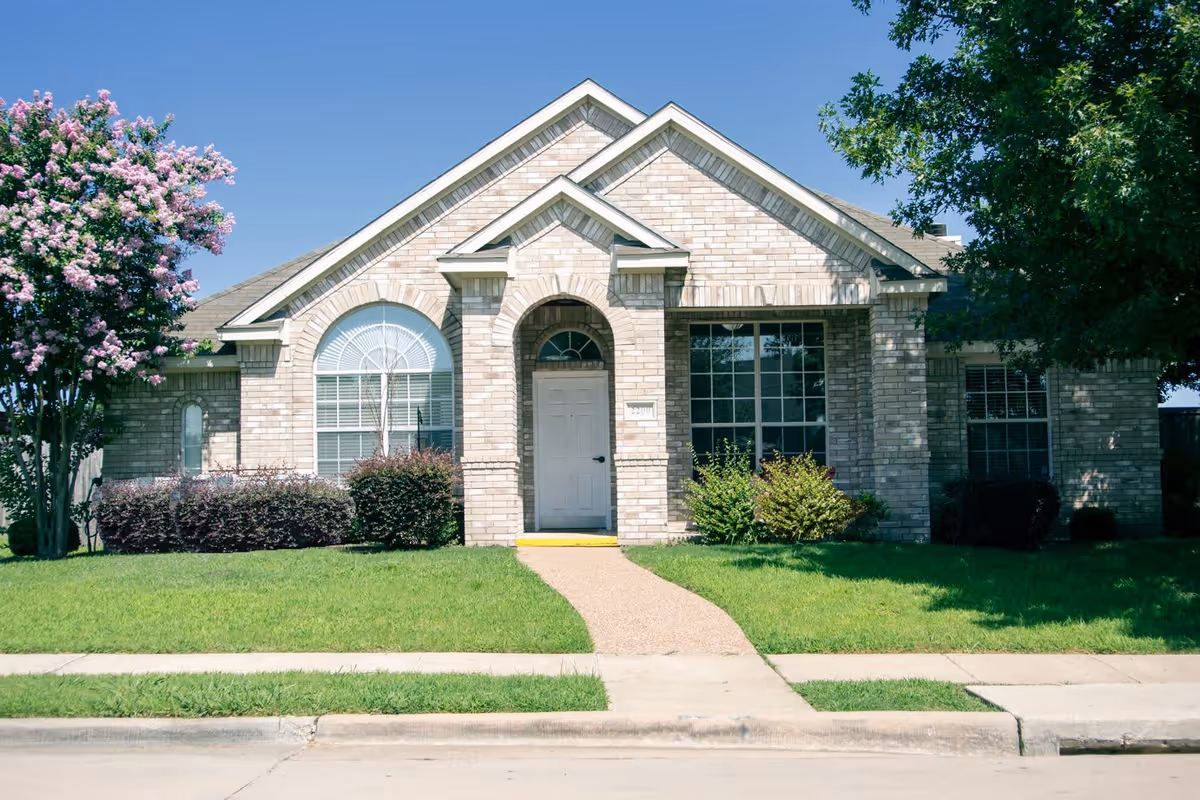 Front exterior view of a single-story brick building with a white door, large windows, and a well-maintained lawn with bushes and trees on either side.