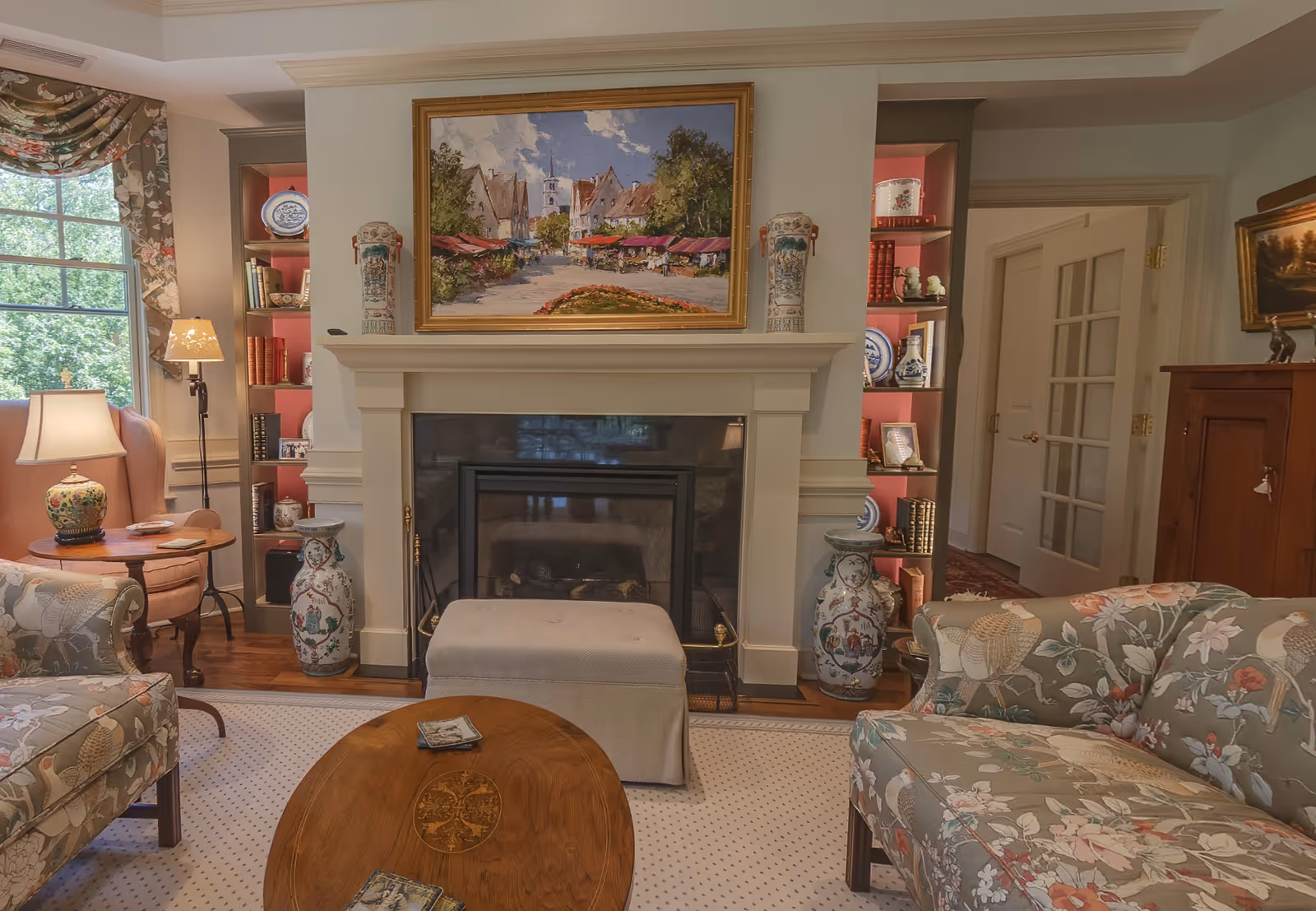 A cozy living room with floral upholstered armchairs and a wooden coffee table. There is a white fireplace with a painting of a village scene above it, flanked by built-in shelves displaying decorative vases, books, and plates. A window with floral curtains lets in natural light, and there are two table lamps providing additional lighting.