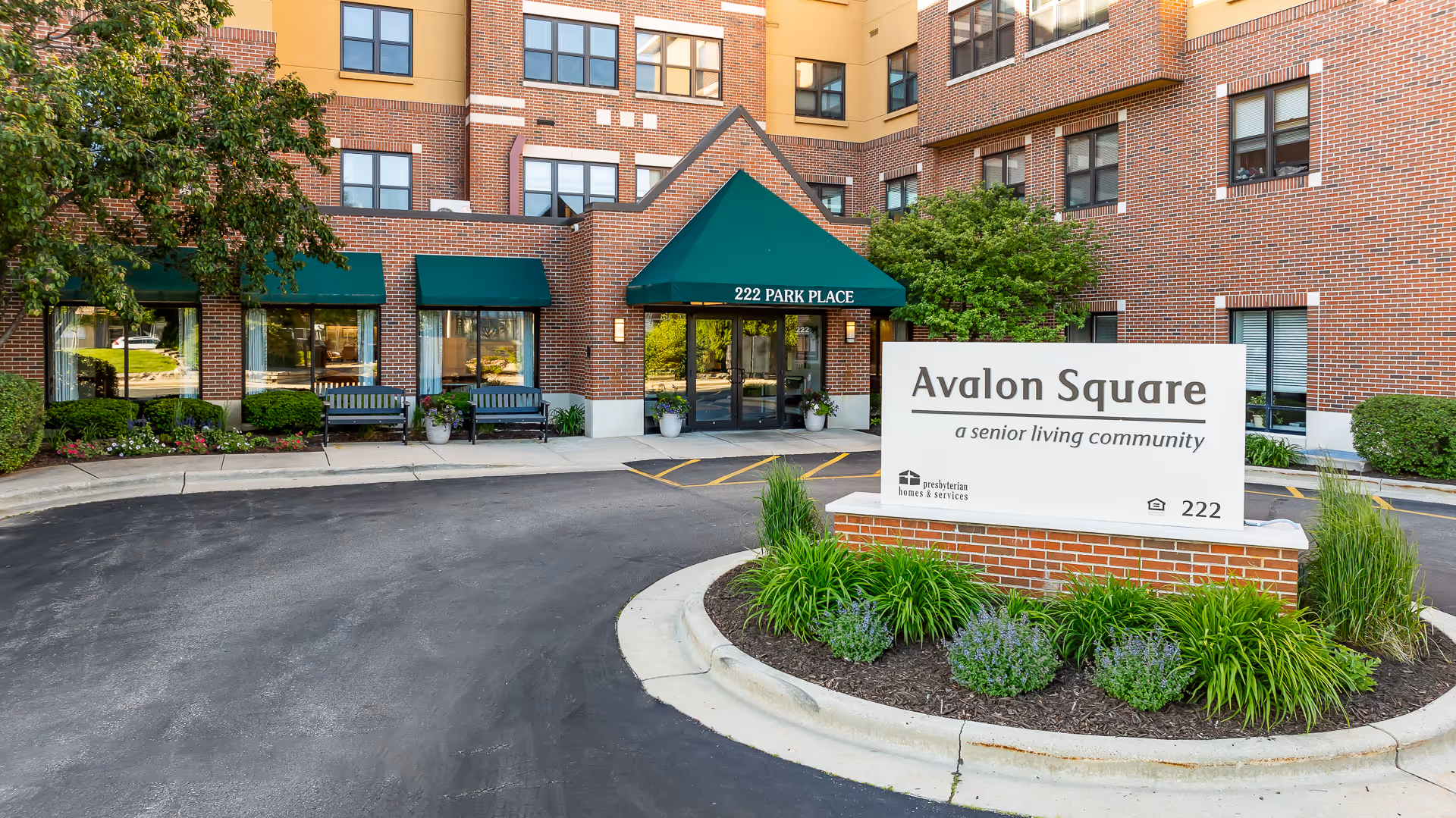 Exterior view of Avalon Square senior living community building with a brick facade, green awnings, benches near the entrance, and a sign displaying the community name and address 222 Park Place surrounded by landscaping.