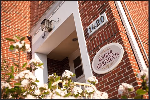 Close-up view of the entrance to a brick building with a white-framed doorway, a lantern-style light fixture above, and a sign that reads 'ROHRER APARTMENTS' along with the building number 1420. White flowers are visible in the foreground.