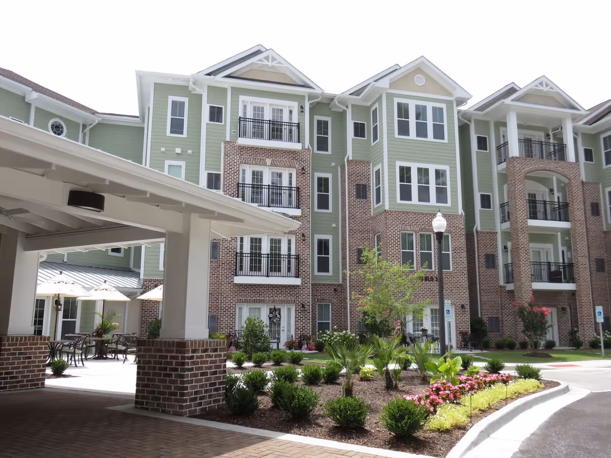 Exterior view of a multi-story senior living facility building with green siding and brick accents. The building features balconies, large windows, landscaped garden beds with shrubs and flowers, a covered entrance area, outdoor seating with umbrellas, and a street lamp.