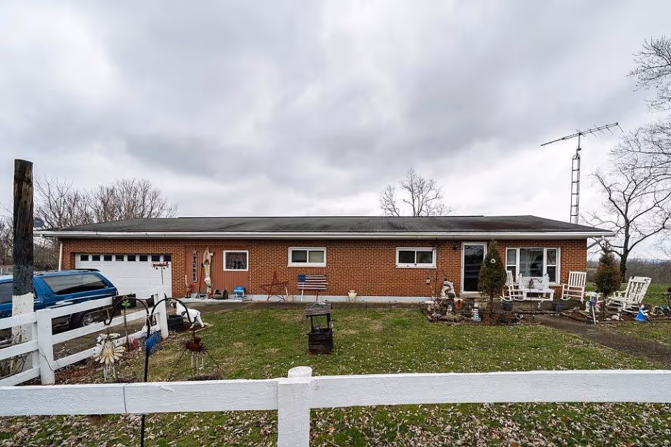 Single-story brick building with a garage on the left side, a front yard with grass and garden decorations, white wooden fence in the foreground, and several white chairs and small trees near the entrance on the right side under a cloudy sky.
