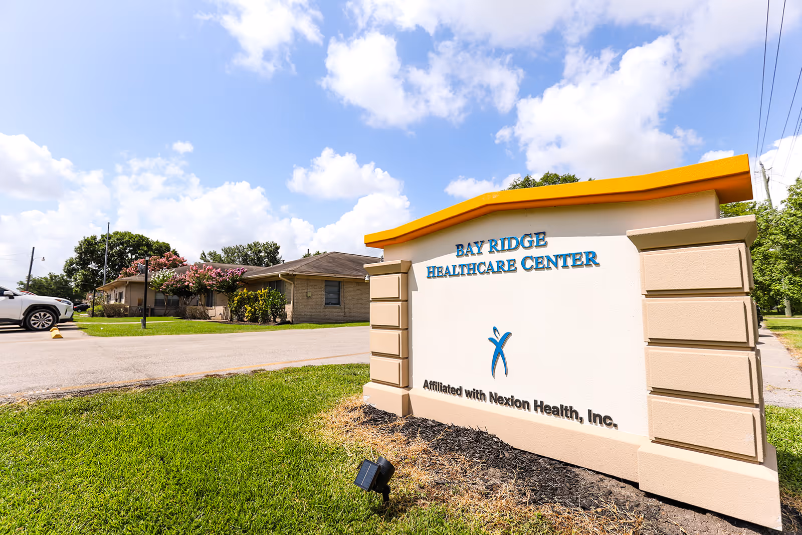 Exterior view of Bay Ridge Healthcare Center sign with a building and parked car in the background under a partly cloudy sky.