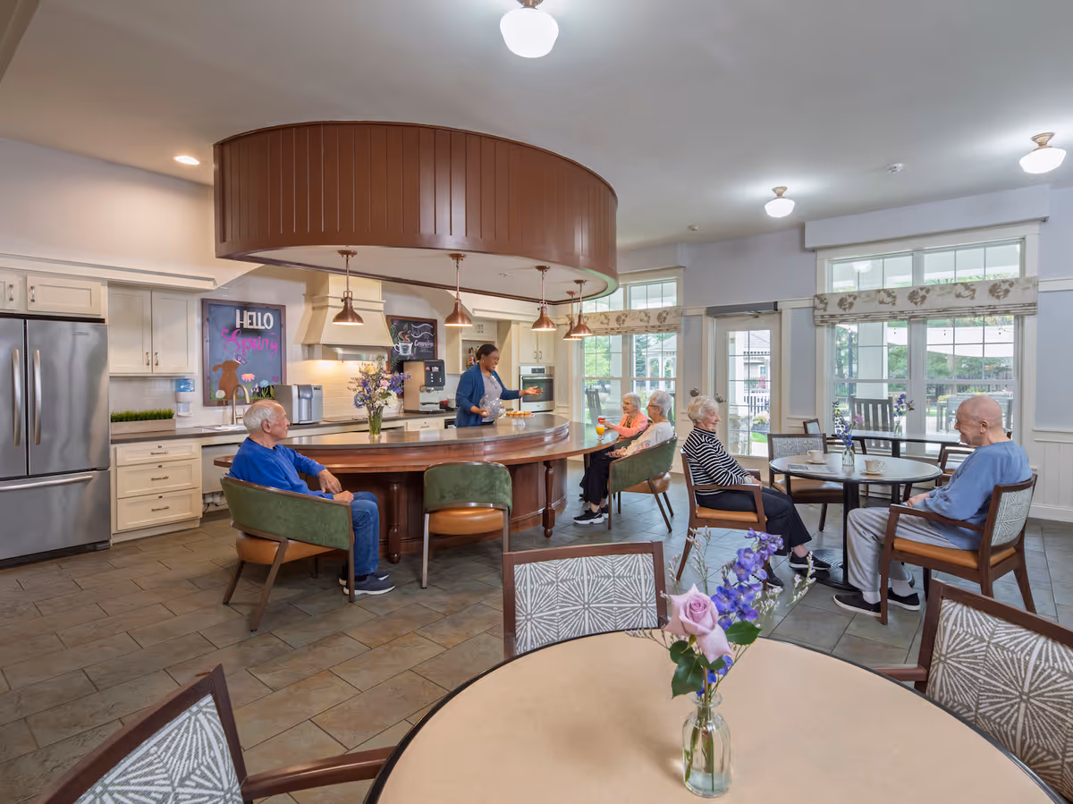 A bright and spacious kitchen and dining area in a senior living facility. Several elderly residents are seated around tables and a curved kitchen island, engaging in conversation. A staff member is serving drinks behind the island. The room features large windows with floral valances, stainless steel appliances, and pendant lighting above the island. Fresh flowers in vases decorate the tables.