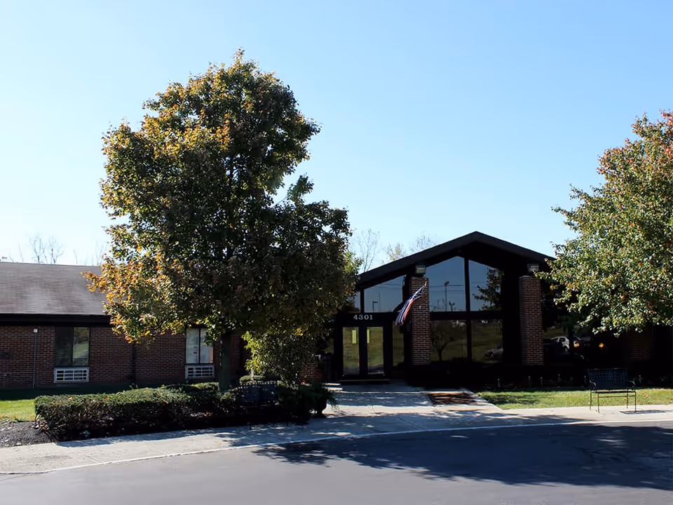 Front exterior view of Columbus Healthcare Center building with a large tree and bushes in front, a paved driveway, and an American flag near the entrance under a clear blue sky.