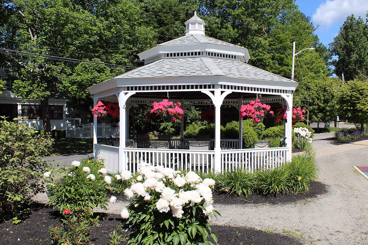 A white wooden gazebo with a shingled roof surrounded by greenery and blooming flowers, including white and pink blossoms. The gazebo has hanging baskets with bright pink flowers and several chairs inside. Trees and a pathway are visible in the background under a clear blue sky.