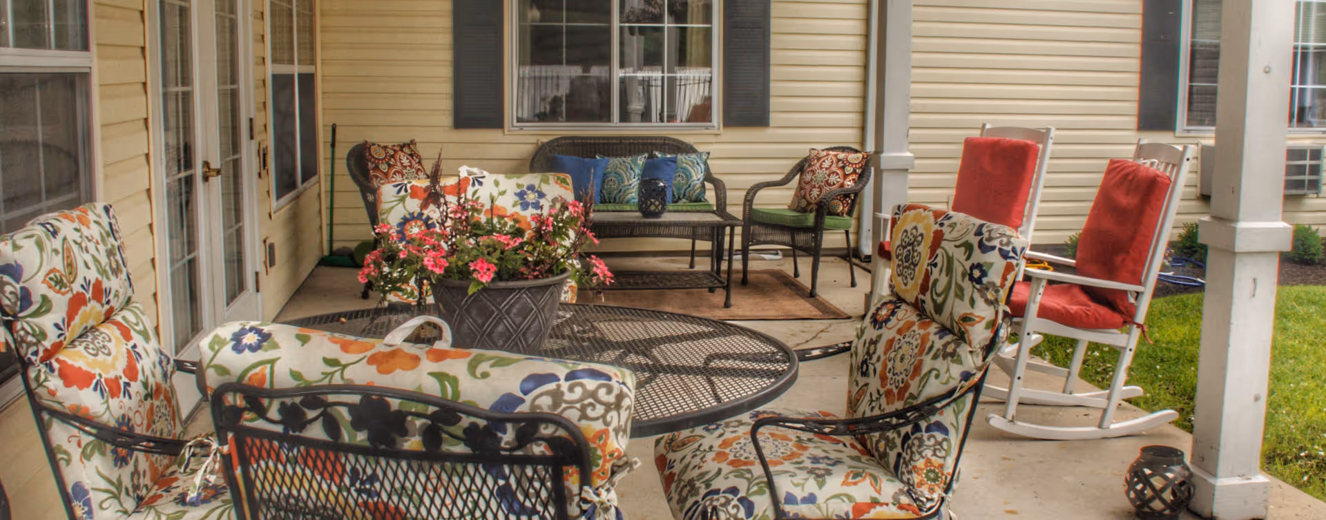 Covered outdoor patio area with metal chairs featuring floral cushions, a round metal table, a flower pot with pink flowers, a wicker loveseat with blue and green cushions, two white rocking chairs with red cushions, and a grassy yard visible beyond the patio.