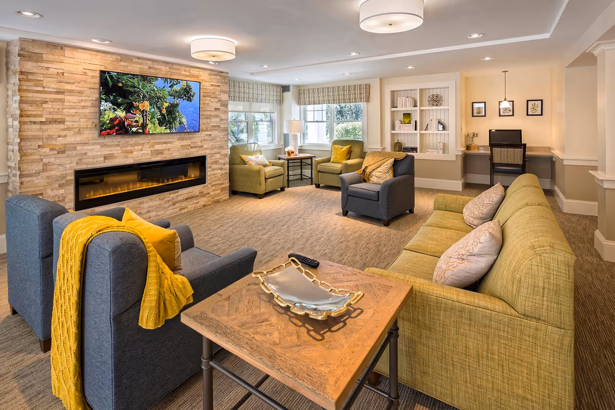 A cozy living room with a stone accent wall featuring a mounted flat-screen TV above a modern electric fireplace. The room has a green sofa with decorative pillows, two green armchairs, and a gray armchair with a yellow throw blanket. A wooden coffee table with a decorative tray is in the foreground. There is a built-in bookshelf with books and decor, a desk with a computer, and large windows letting in natural light.