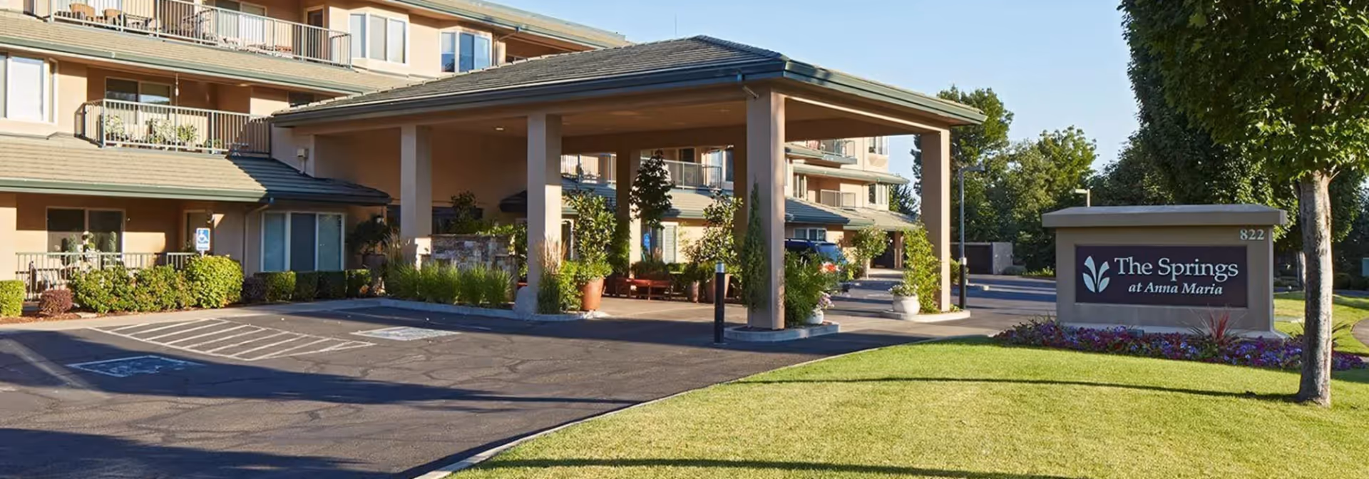 Exterior view of The Springs at Anna Maria senior living facility showing the entrance with a covered drop-off area, surrounding greenery, and a sign with the facility name.