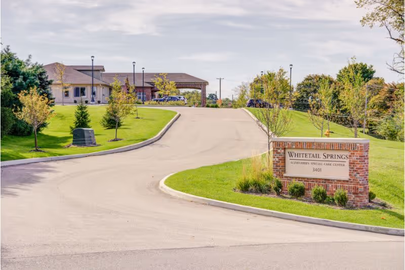 View of the entrance driveway leading up to Whitetail Springs Memory Care Community building, with a brick sign on the right side displaying the facility name and address, surrounded by green grass, small trees, and landscaping under a partly cloudy sky.