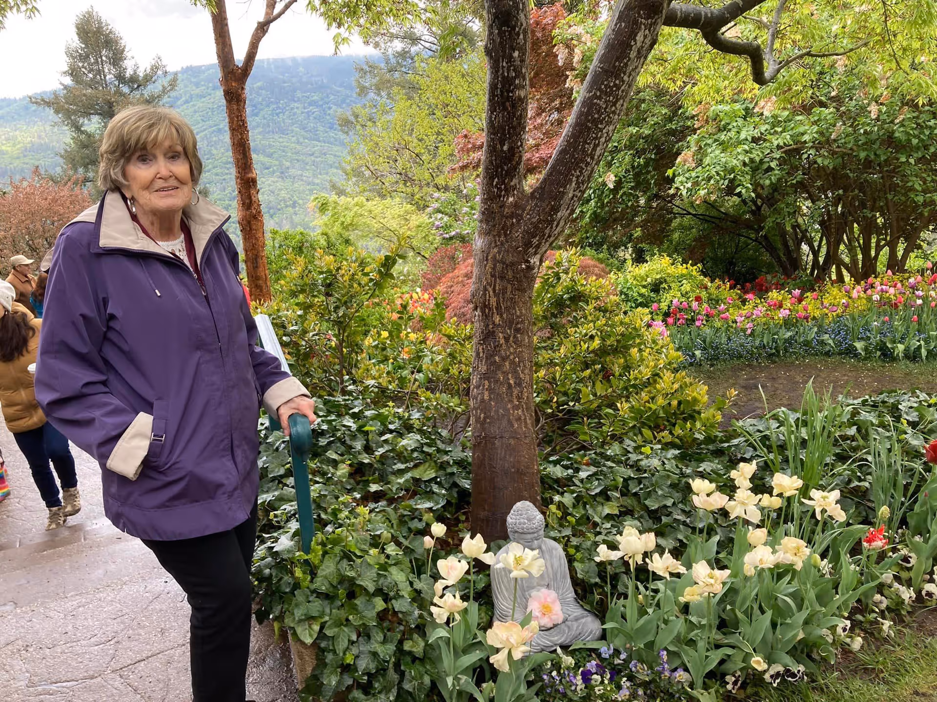 An elderly woman wearing a purple jacket stands on a garden path next to a green handrail. She is surrounded by lush greenery, colorful flowers, and a small Buddha statue near the base of a tree. In the background, there are other people walking and a view of forested hills under a cloudy sky.