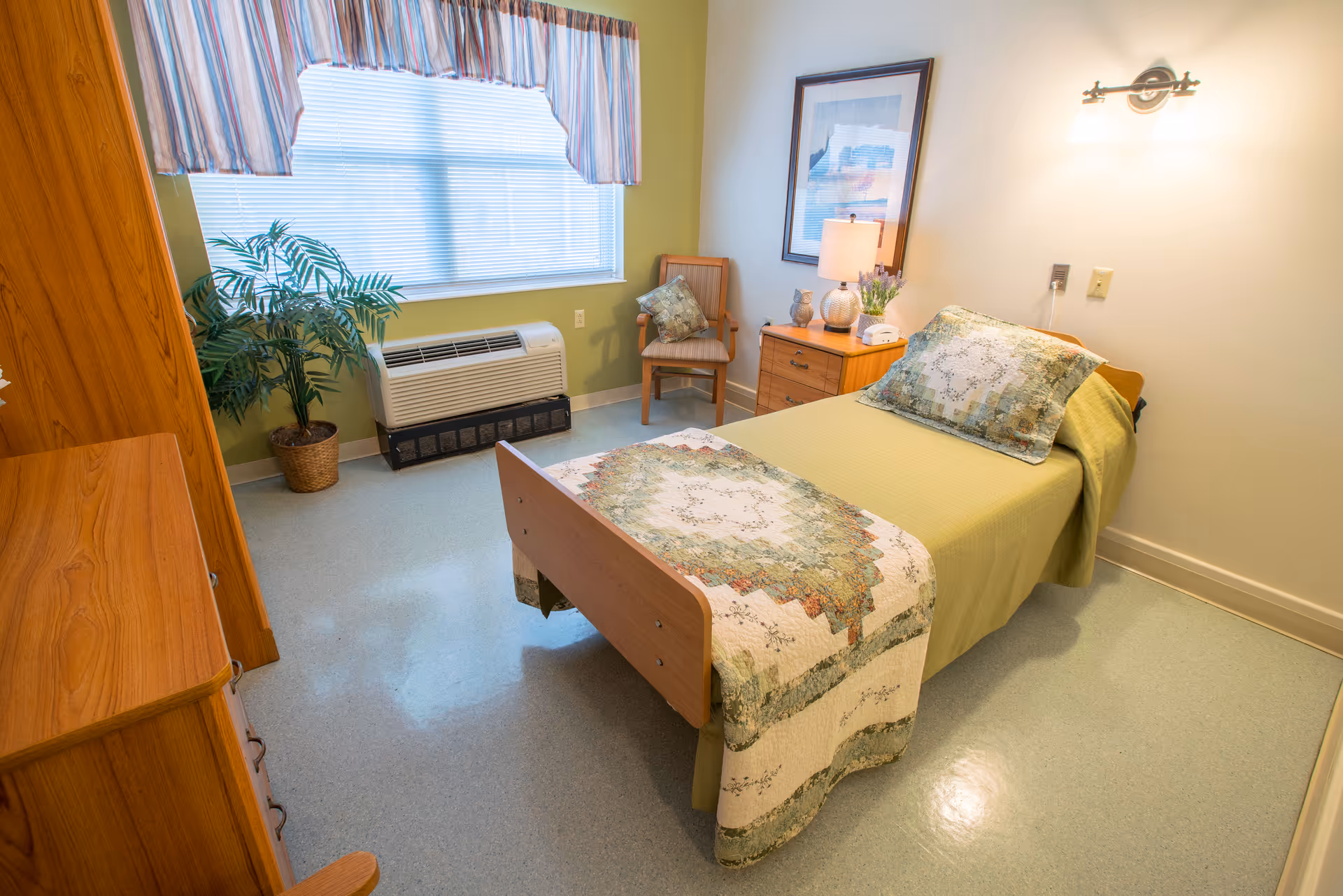 A small, neatly arranged bedroom in a senior living facility with a single bed covered in a green bedspread and a decorative quilt. The room has a wooden nightstand with a lamp, a chair with a cushion, a framed picture on the wall, a large window with striped curtains, a potted plant, and a heating/cooling unit beneath the window.