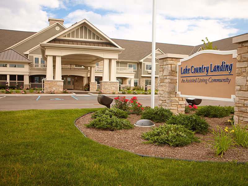 Exterior view of Lake Country Landing assisted living community building with a covered entrance, landscaped garden with bushes and flowers, and a sign displaying the facility name.