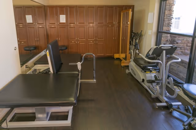 A small fitness or physical therapy room with a padded treatment table, exercise equipment including a recumbent bike, a full-length mirror, and wooden cabinets along the back wall. There is a large window letting in natural light.