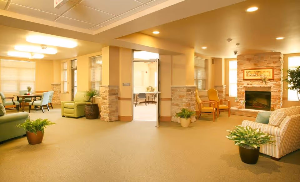 A spacious and warmly lit living room area in a senior living facility with comfortable armchairs, a striped sofa, potted plants, and a stone fireplace with a framed picture above it. There is a doorway leading to another room with tables and chairs visible.